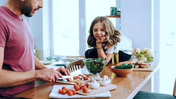 Ein Mann und ein Mädchen bereiten zusammen einen Salat zu. Der Mann schneidet Tomaten und Pilze auf einem Brett, das Mädchen sieht ihm dabei zu. Sie unterhalten sich dabei. Auf dem Tisch stehen außerdem eine Salatschüssel mit etwas Salat, geschnittenes Brot sowie Gurken, Möhren und Äpfel in einem Obstkorb.