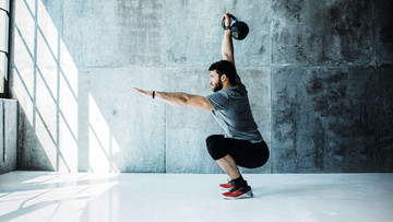 Ein Mann steht in einer kargen Fitnesshalle vor grauer Betonwand in einer tief gebeugten Squat-Position. Sein linker Armer ist nach vorn ausgestreckt, mit dem rechten stemmt er ein Gewicht nach oben.