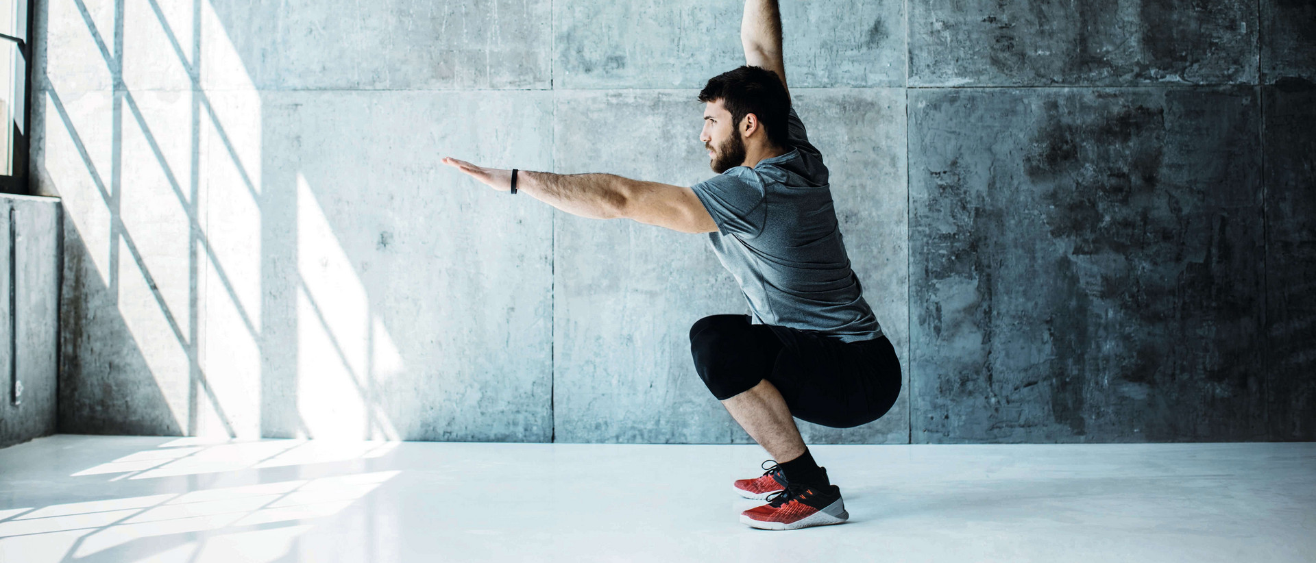 Ein Mann steht in einer kargen Fitnesshalle vor grauer Betonwand in einer tief gebeugten Squat-Position. Sein linker Armer ist nach vorn ausgestreckt, mit dem rechten stemmt er ein Gewicht nach oben.