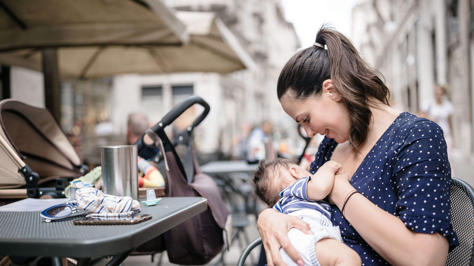 Eine Mutter mit dunklen Haaren – zu einem Zopf gebunden – stillt ihren kleinen Sohn. Das Baby hat dunkle Haare und trägt ein T-Shirt und Windel. Sie sitzen in einem Restaurant an einem Tisch mitten in einer Fußgängerzone. 