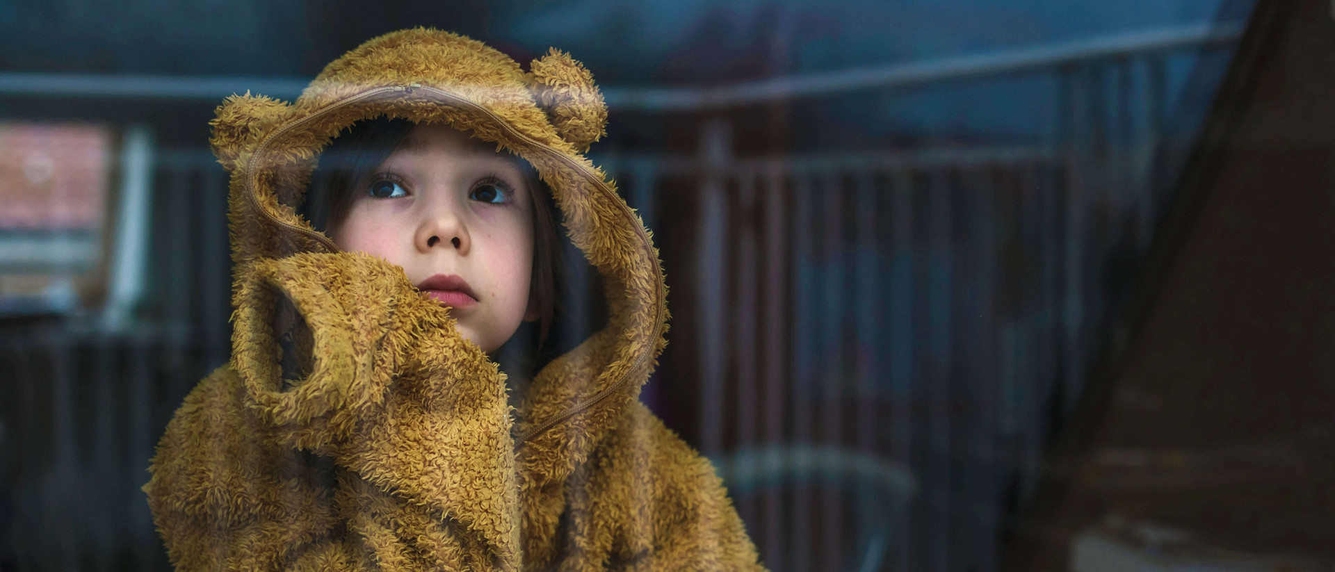 Ein Kind mit braunen Haaren blickt traurig in die Ferne. Es trägt ein Bärenkostüm und wirkt einsam. 