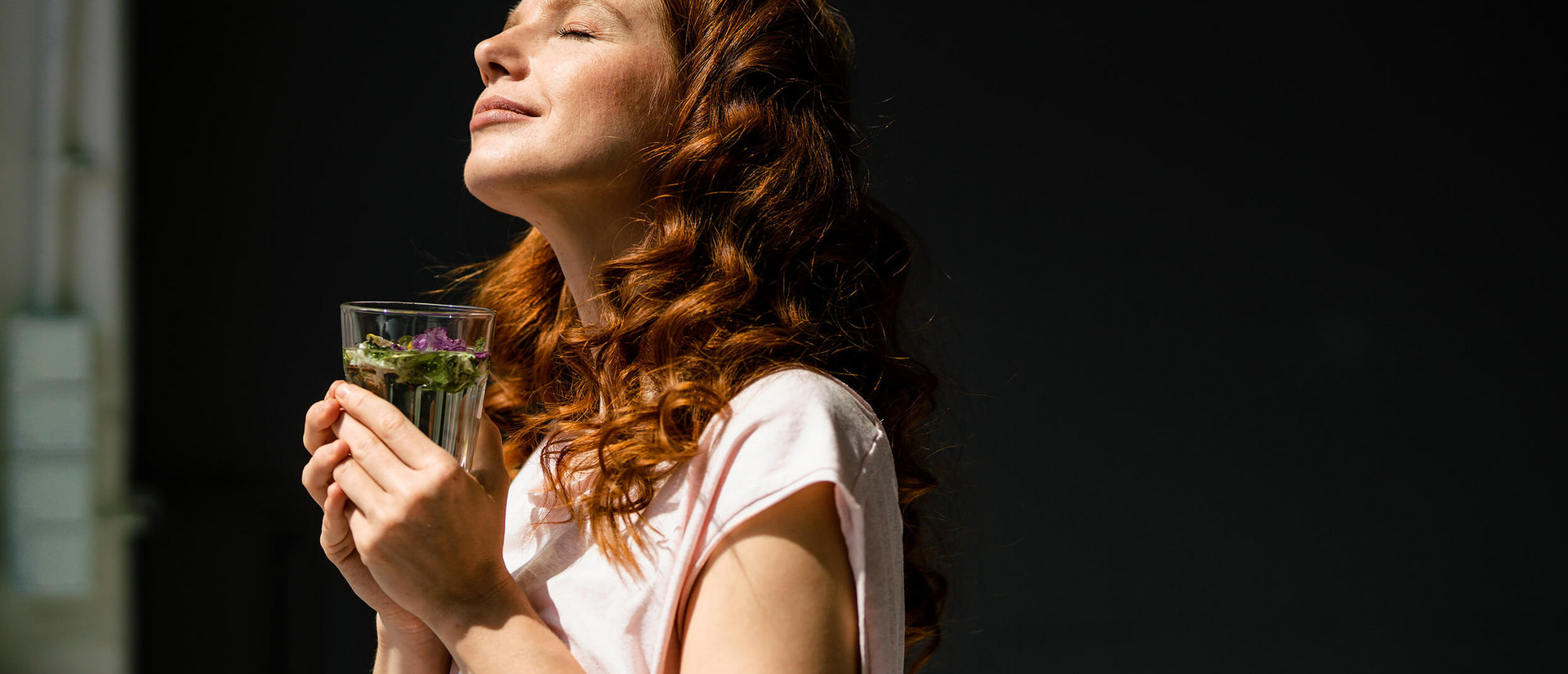 Eine junge, rothaarige Frau steht mit einem Glas Tee vor dem Fenster. Sie streckt ihr Gesicht mit geschlossenen Augen der Sonne entgegen.
