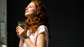 Eine junge, rothaarige Frau steht mit einem Glas Tee vor dem Fenster. Sie streckt ihr Gesicht mit geschlossenen Augen der Sonne entgegen.
