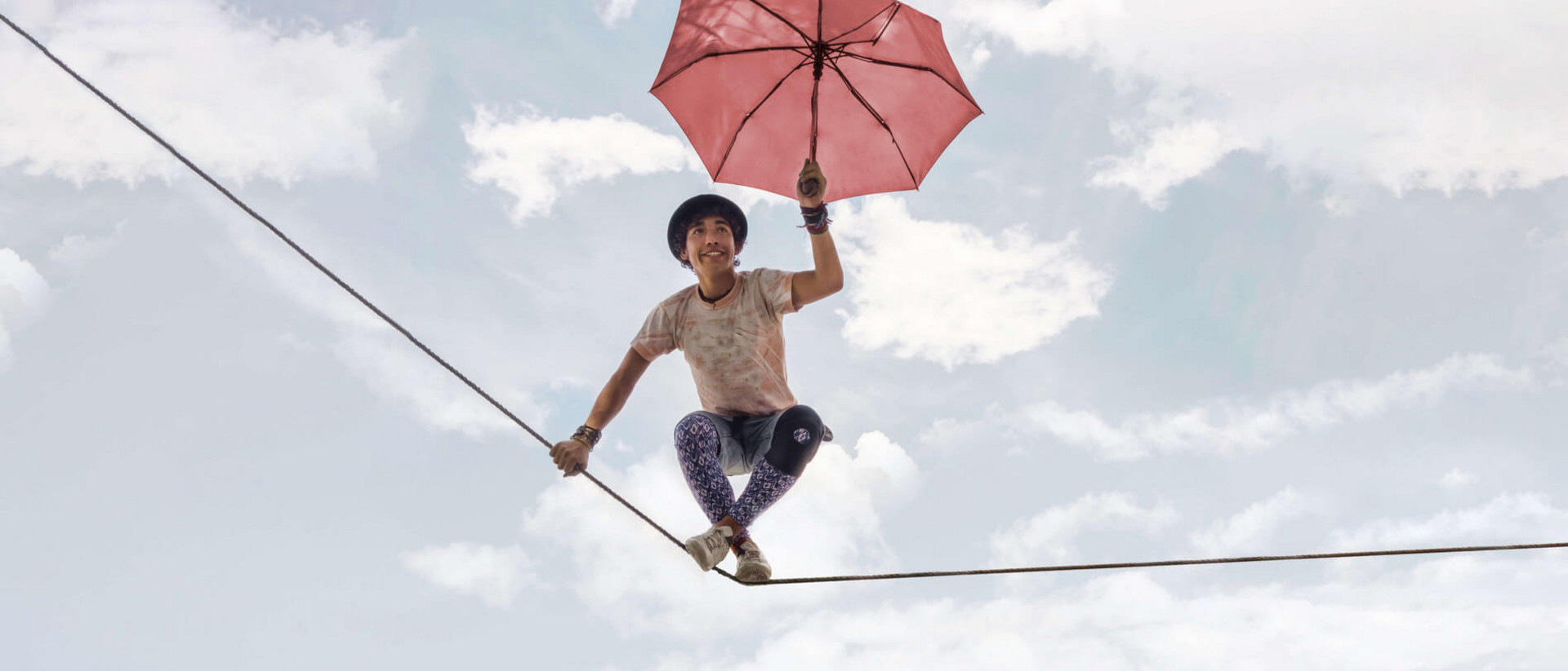 Ein junger Mann balanciert fröhlich auf einem Seil. In der Hand hält er einen geöffneten Regenschirm.