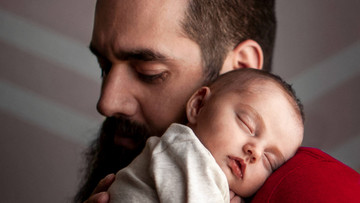Ein junger Mann hat ein schlafendes Baby auf seiner Schulter liegen. Der Mann hält ihm fürsorglich den Rücken.