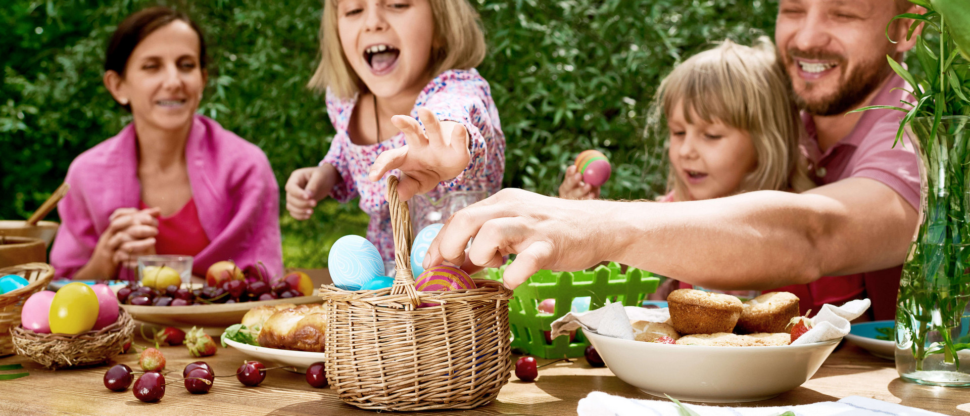 Ein Mann, eine Frau und zwei Mädchen sitzen an einem Holztisch. Der Mann und eines der Mädchen greifen nach bunt angemalten Eiern, die in einem Körbchen auf dem Tisch stehen. Das zweite Mädchen hält ebenfalls ein bunt angemaltes Ei in der Hand, die Frau schaut zu und lacht. 