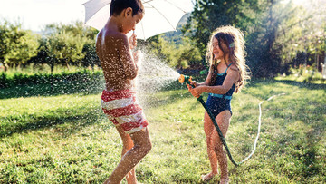Ein Junge und ein Mädchen spielen auf einer Wiese mit einem Wasserschlauch. Das Mädchen spritzt den Jungen nass und lacht dabei, der Junge hält einen kleinen weißen Schirm in der Hand. Beide tragen Badekleidung. 