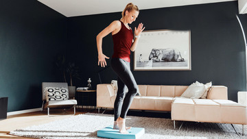 Die Sportwissenschaftlerin Ingalena Schömburg-Heuck steht in Sportkleidung in einem Wohnzimmer. Sie macht eine Balanceübung auf einem Balance-Pad vor.