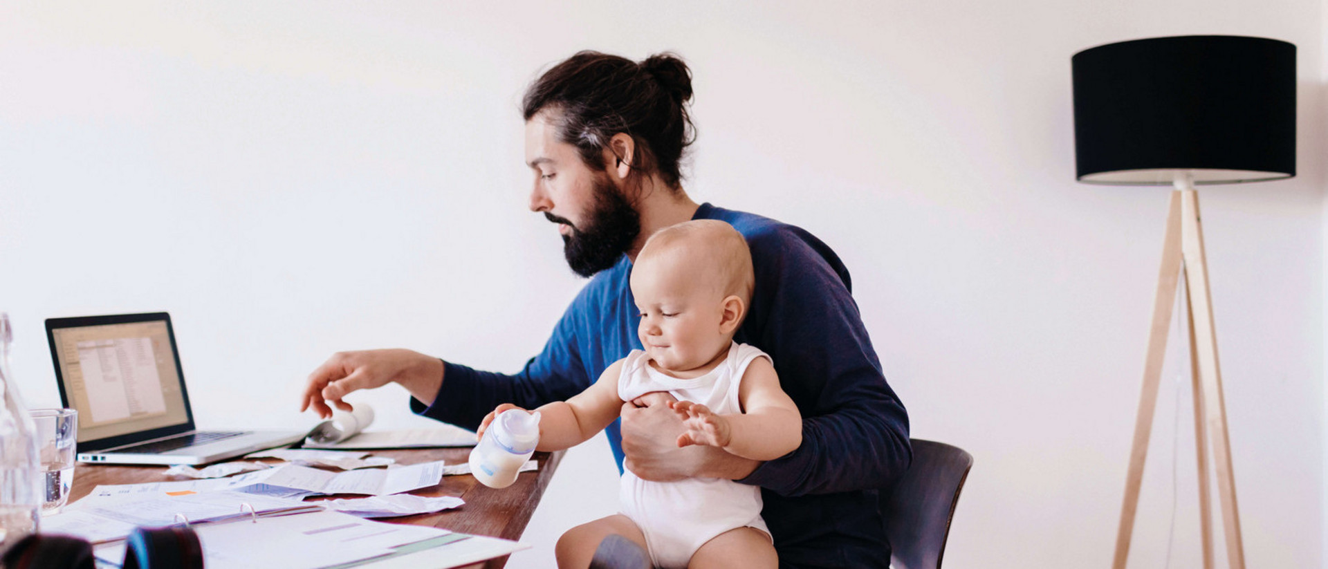 Ein Mann sitzt vor seinem Computer am Schreibtisch. Auf seinem Schoß sitzt ein Baby, das mit einer Babyflasche spielt. 