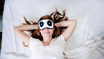 Junge Frau trägt eine Schlafmaske mit Panda-Augen und liegt mit verschränkten Armen hinter dem Kopf im Bett