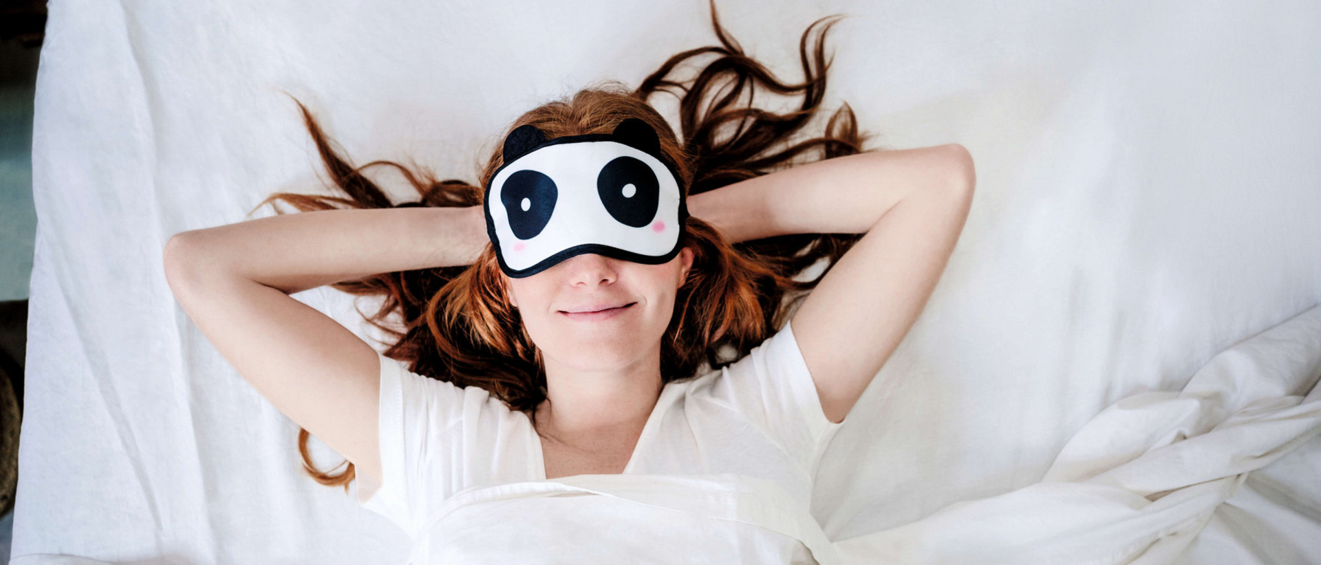 Junge Frau trägt eine Schlafmaske mit Panda-Augen und liegt mit verschränkten Armen hinter dem Kopf im Bett