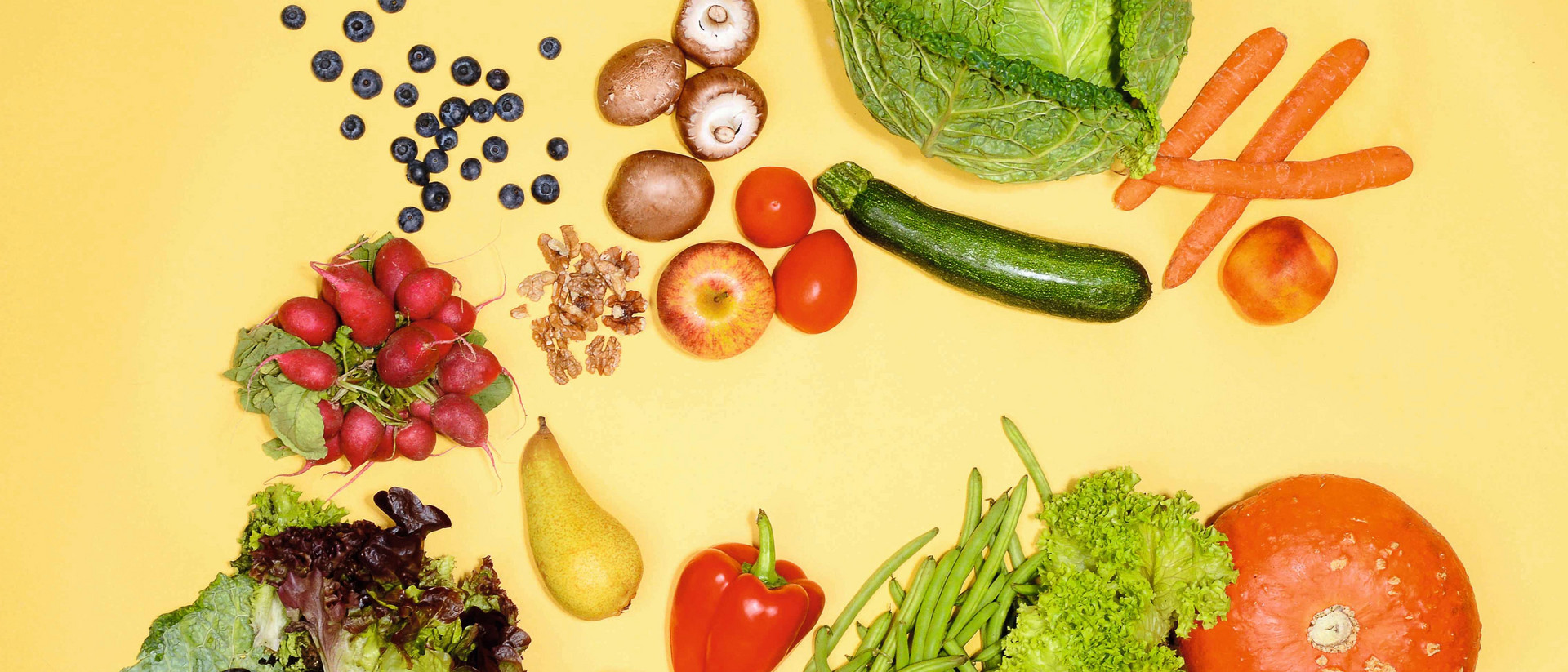 Gemüse und Obst unterschiedlicher Art liegen lose verstreut auf einem gelben Untergrund.