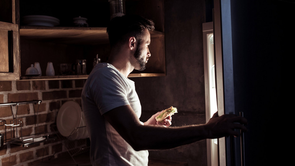 Junger Mann steht im Pyjama im dunklen vor der geöffneten Kühlschranktüre