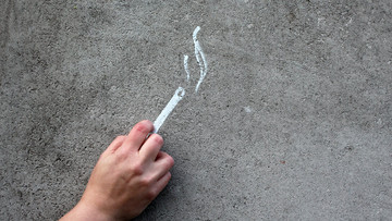 Auf einen grauen Asphaltboden ist eine weiße Zigarette gemalt. Eine Hand liegt auf dem Asphaltboden auf der Höhe der Zigarette, sodass es aussieht, als ob sie sie hält. 