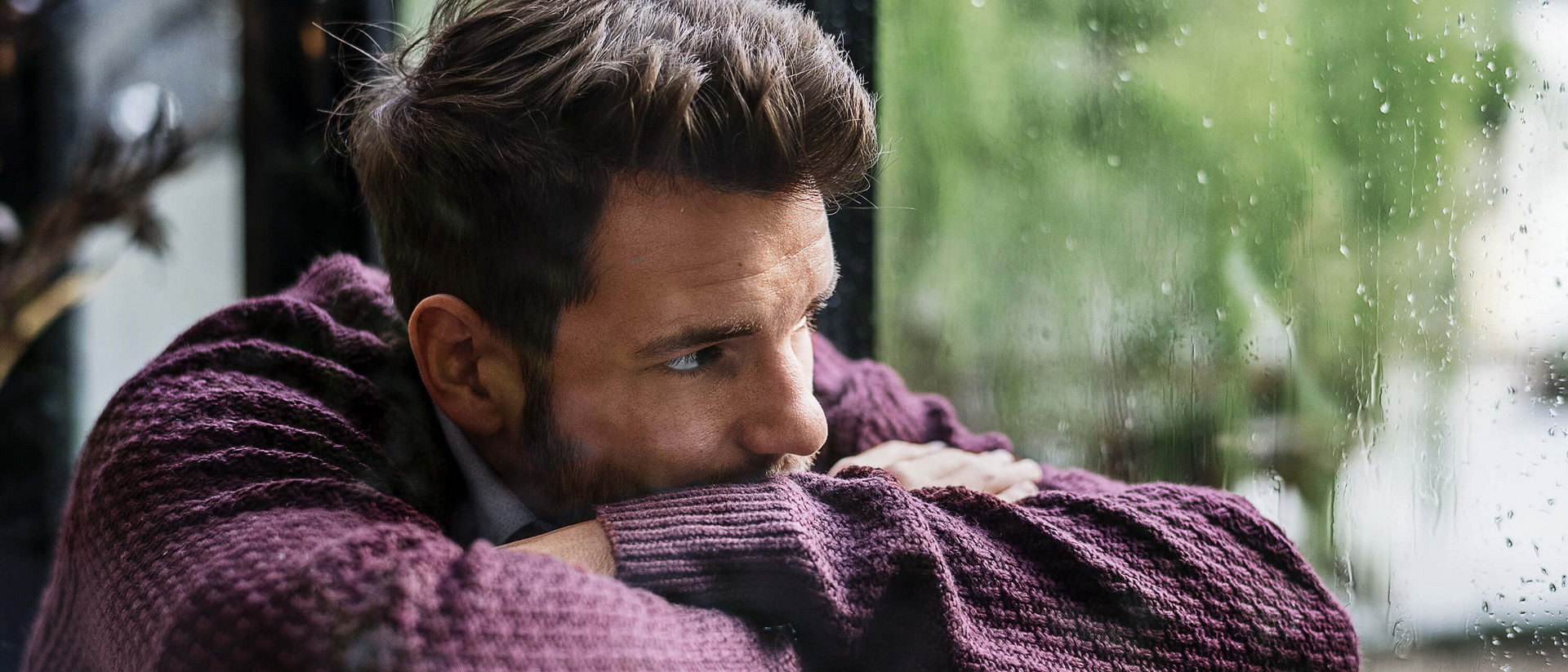 Ein junger Mann hat den Kopf auf die verschränkten Arme gelegt und schaut gedankenverloren durch ein regennasses Fenster in die Ferne.