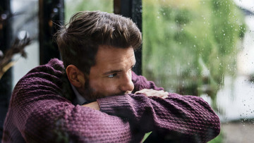 Ein junger Mann hat den Kopf auf die verschränkten Arme gelegt und schaut gedankenverloren durch ein regennasses Fenster in die Ferne.