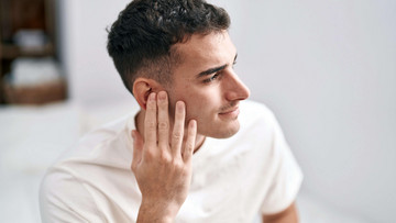 Ein junger Mann mit braunen Locken ist im Profil zu sehen. Er hält seine Hand an sein rechtes Ohr.