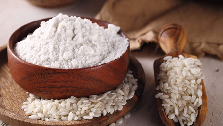 Ein braunes Gefäß mit glutenfreiem Reismehl steht auf einem braunen Untersetzer. Daneben ist eine kleine Portion Reis zu sehen. 