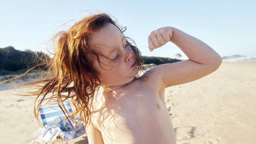 Ein kleines Kind mit wilden, roten langen Haaren hält den linken Arm angewinkelt nach oben und spannt den Bizeps an. Im Hintergrund sind ein Strand, ein Stuhl und Dünen zu sehen. Das Kind hat einen freien Oberkörper. 