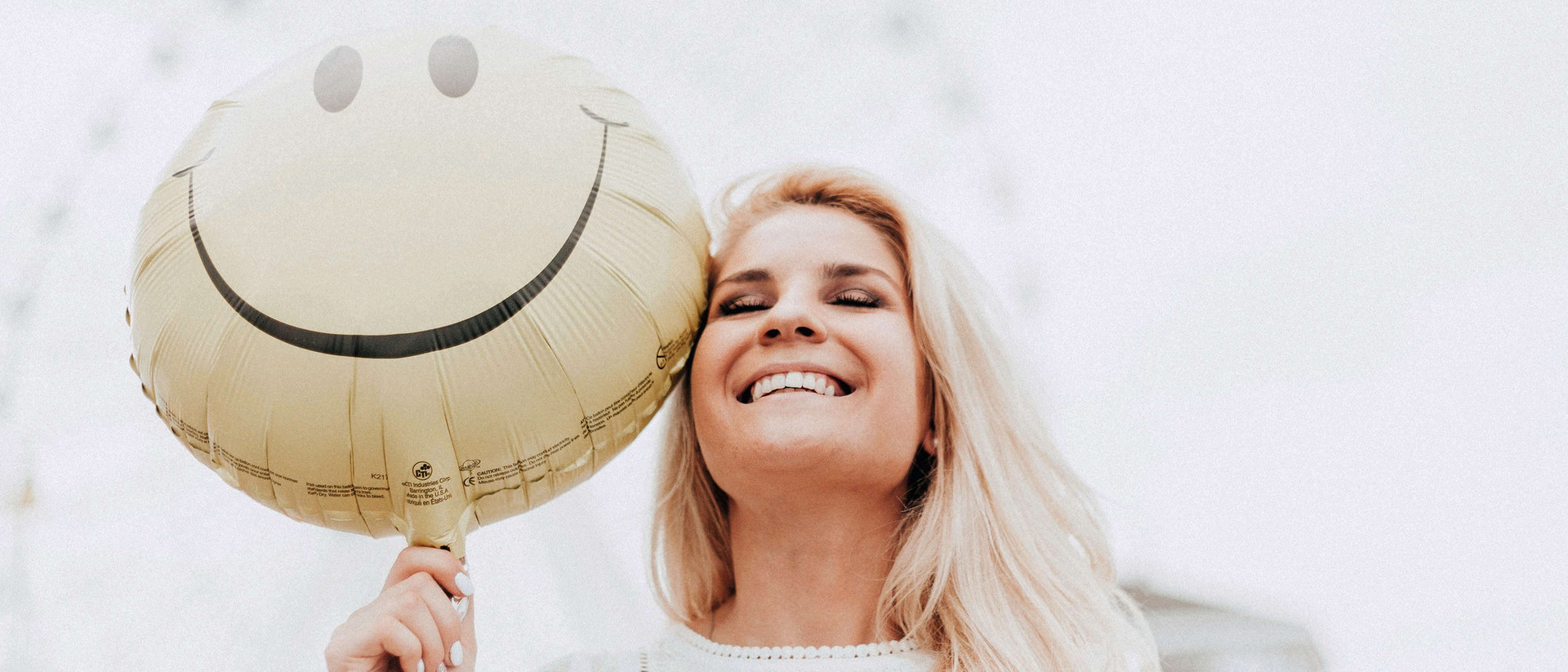 Eine blonde Frau lacht mit geschlossen Augen. Zu ihrer Linken, direkt neben ihrem Gesicht, ist ein Luftballon zu sehen, auf dem ein lachendes Gesicht zu sehen ist.