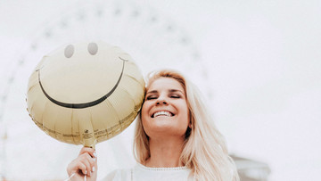 Eine blonde Frau lacht mit geschlossen Augen. Zu ihrer Linken, direkt neben ihrem Gesicht, ist ein Luftballon zu sehen, auf dem ein lachendes Gesicht zu sehen ist.