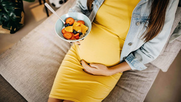 Eine schwangere Frau sitze auf der Couch. Mit der einen Hand streichet sie über ihren kugelrunden Bauch, in der anderen Hand hält sie eine Schale mit Obstsalat oder Müsli