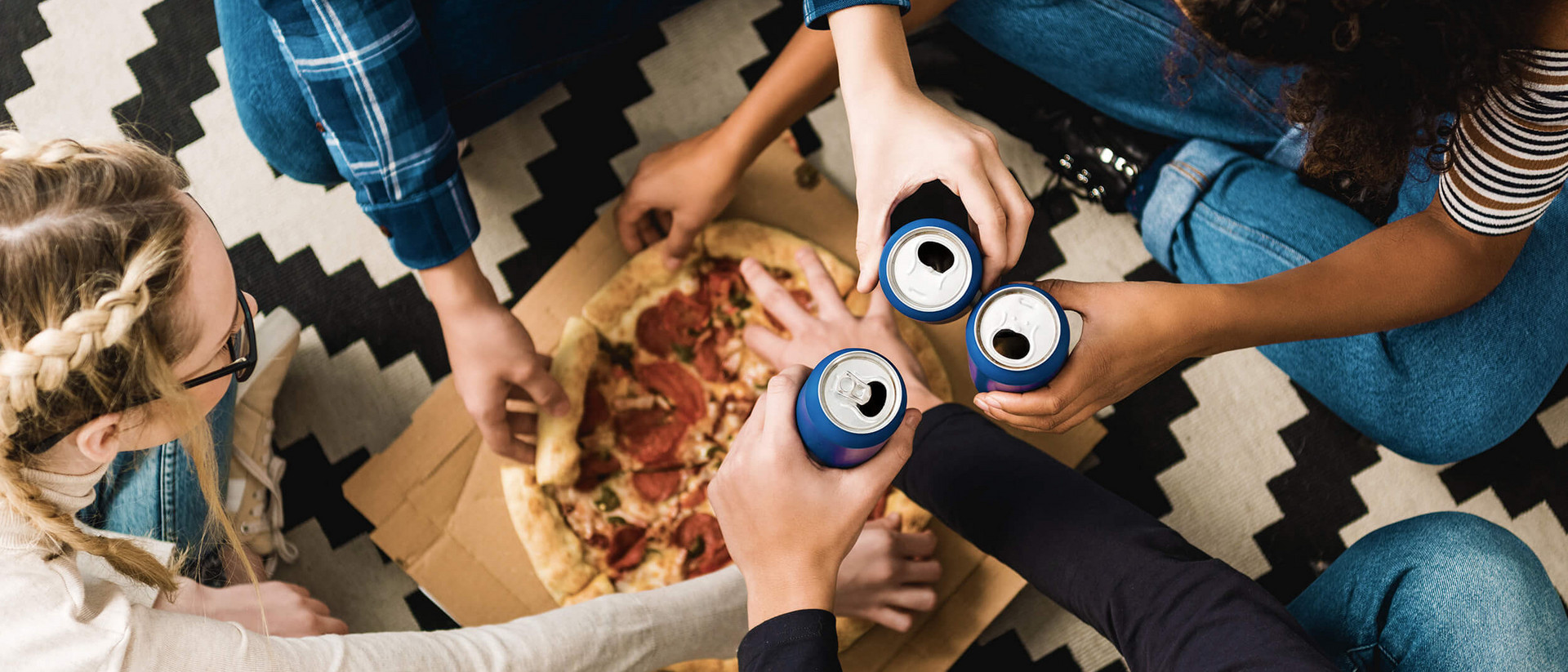 Vier junge Menschen sitzen im Schneidersitz auf dem Boden. In der Mitte von ihnen liegt eine Pizza auf einem Pappkarton. Alle vier nehmen sich ein Stück Pizza, drei von ihnen halten dabei eine blaue Dose in der anderen Hand und stoßen damit an. 