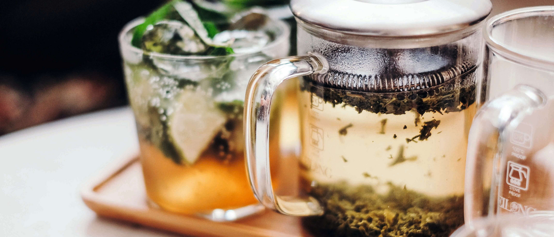 Eine gläserne Teekanne steht auf einem kleinen braunen Tablett, links und rechts von ihr stehen gläserne Teetassen. Die Kanne ist mit Wasser und frischem Tee gefüllt, das Glas links mit Tee und frischer Zitrone, das Glas rechts ist leer. 