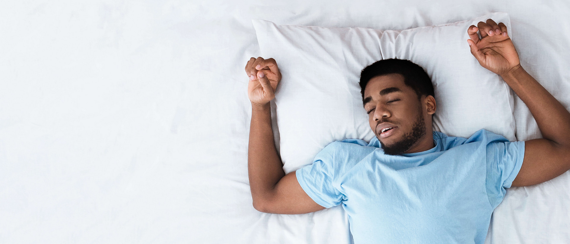 Mann liegt schlafend im Bett, die Arme sind beidseitig nach oben angewinkelt, als würde er Gewichte stemmen.