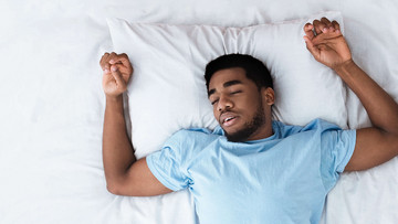 Mann liegt schlafend im Bett, die Arme sind beidseitig nach oben angewinkelt, als würde er Gewichte stemmen.