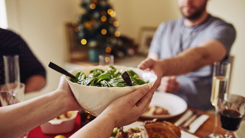An einem festlich gedeckten Tisch reicht jemand eine Schüssel mit grünem Salat an den Tischnachbarn weiter. Im Hintergrund steht ein Weihnachtsbaum.