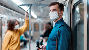 Ein Mann steht in einer U-Bahn, trägt eine Maske und hält ein Handy in der Hand. 