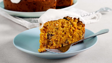 Ein Stück orangefarbener Kuchen mit weißem Topping liegt auf einem türkisfarbenen Teller, daneben eine Gabel. Der Teller steht auf einem grauen Tisch. Im Hintergrund ist der übrige Kuchen – ebenfalls auf einem türkisfarbenen Teller, zu sehen. 