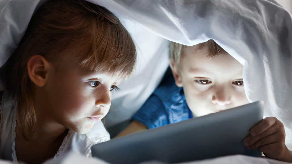 Zwei Kinder haben eine Decke über ihre Köpfe gezogen und schauen unter der Decke auf ein Tablet. 