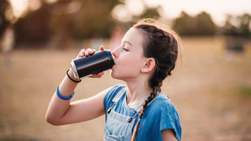 Ein junges Mädchen mit dunkelblonden Haaren, zu zwei Zöpfen gebunden, trinkt aus einer schwarzen Dose. Sie trägt eine Latzhose mit blauen Streifen und ein blaues Oberteil. 
