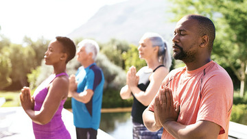 Vier Menschen, zwei Männer und zwei Frauen, haben sich zur Meditation im Freien versammelt. Sie halten beide Handflächen in Brusthöhe aneinander, ihre Augen sind geschlossen. Im Hintergrund ist eine grüne, bergige Landschaft zu sehen.