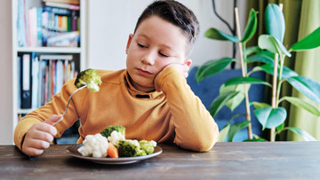 Ein braunhaariger Junge sitzt an einem Tisch. Vor ihm steht ein Teller mit Gemüse: Blumenkohl, Brokkoli und Möhren. In der rechten Hand hält der Junge eine Gabel mit einem Brokkoli-Röschen. Er schaut es missmutig an.