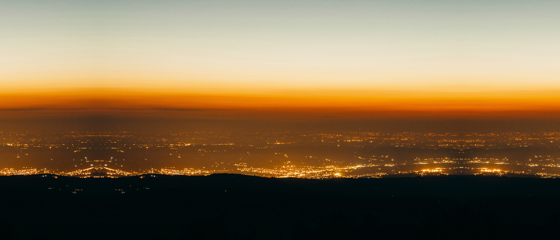 Aufnahme einer Landschaft im Morgengrauen: Alle naheliegenden Objekte versinken noch im Schwarz, während die Lichter der Stadt im Hintergrund orange leuchten.