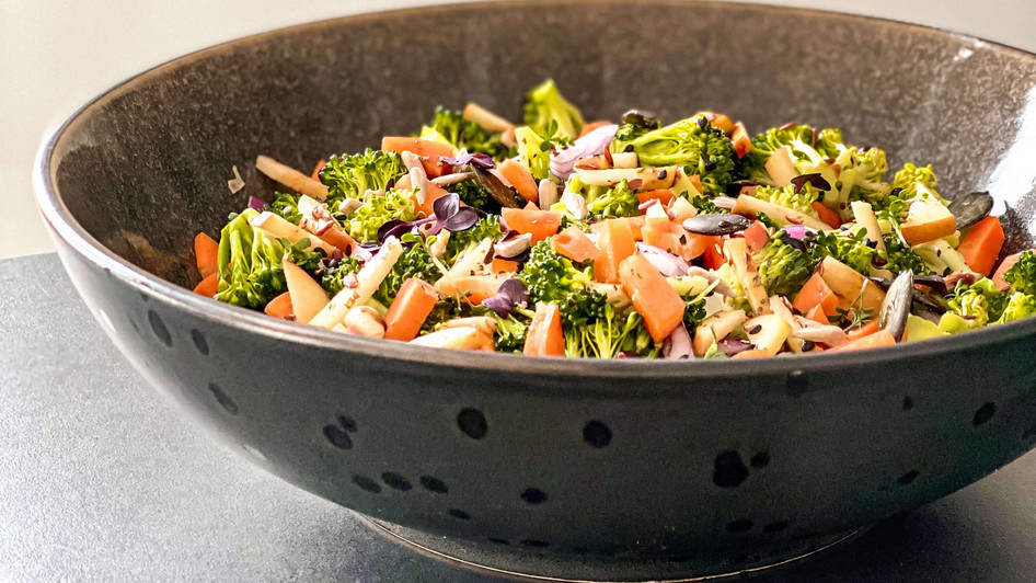 Eine graue Porzellanschale steht auf einem Tisch, sie ist befüllt mit gemischtem Gemüse wie beispielsweise Möhren, Brokkoli und Zwiebeln.