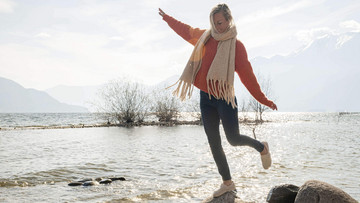 Ja klar! Ein junge blonde Frau balanciert vor einer Bergseekulisse auf einem Bein auf einem Stein.