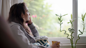 Eine Frau mit dunklen gelockten Haaren sitzt, in eine Decke eingehüllt, an einem Fenster. Sie blickt hinaus, in ihrer rechten Hand hält sie eine Tasse. Rechts von ihr steht eine grüne Pflanze. 