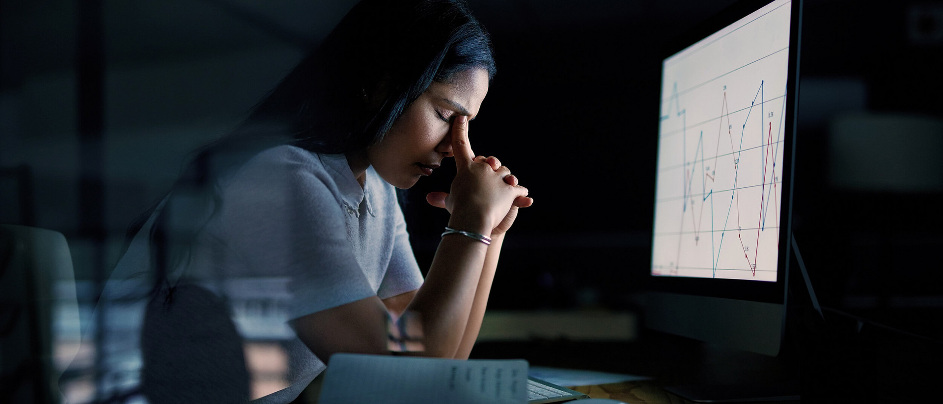 Eine Frau mit dunklen langen Haaren sitzt in einem düsteren Zimmer vor einem hell erleuchteten Computer-Bildschirm. Sie trägt ein weißes T-Shirt und hält ihre gefalteten Hände auf Höhe ihres Gesichts. Mit den Zeigefingern reibt sie sich die Augen. 