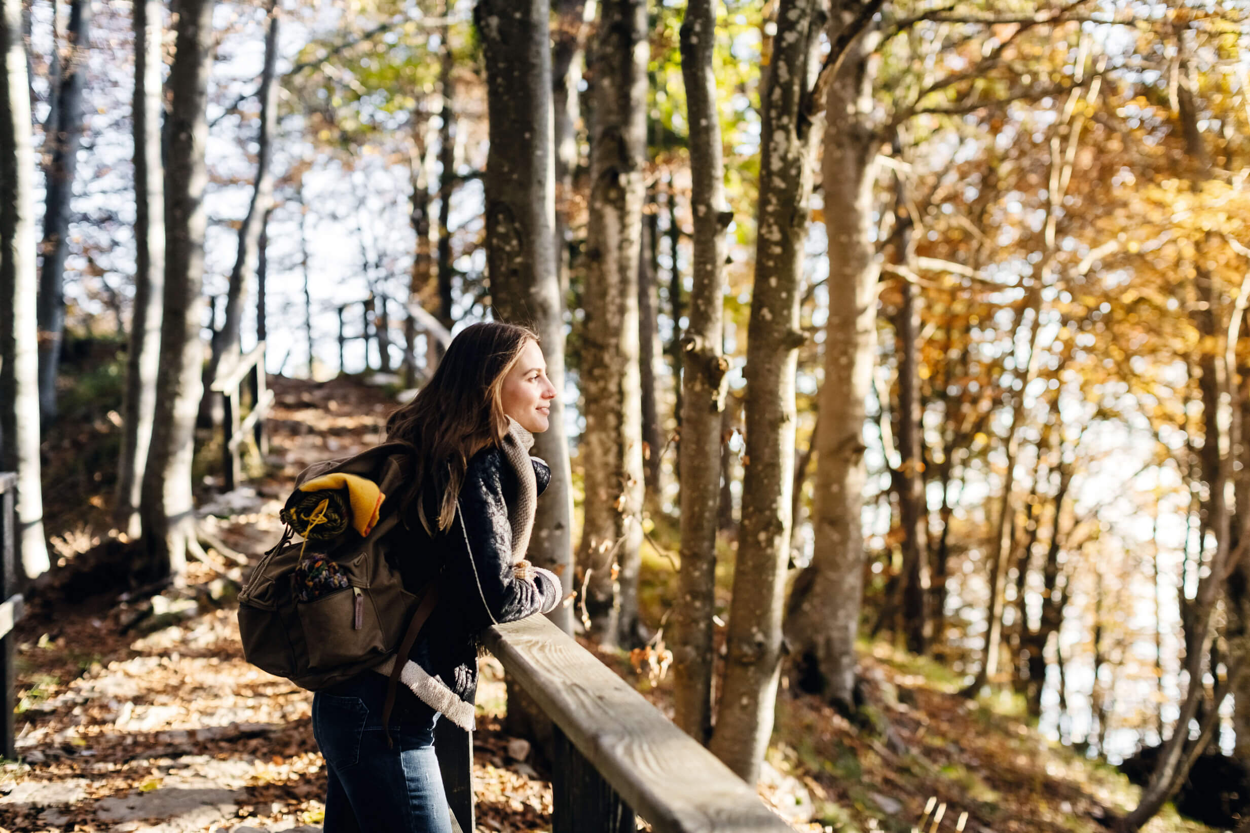 Eine junge Frau lehnt in einem Herbstwald an einem Holzgeländer und schaut lächelnd in die Natur.